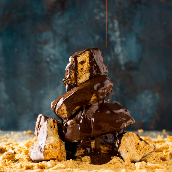 Stack of honeycomb pieces being drizzled with dark chocolate on top, set against a blue textured background.