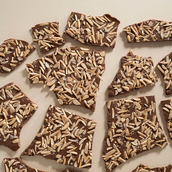 Broken pieces of milk chocolate bark topped with roasted almonds and smoked salt, displayed on a neutral background.